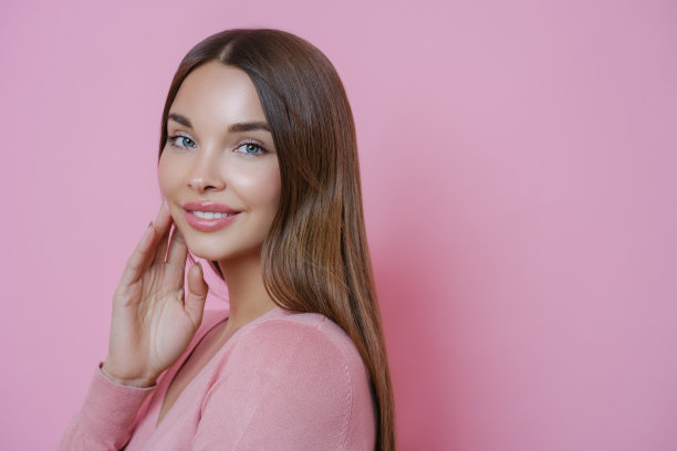 照片中，一位美丽的棕色头发的白人女性把手放在脸颊上，温柔地微笑，直视镜头，蓝眼睛，淡妆，站在粉红色背景的侧面图片下载