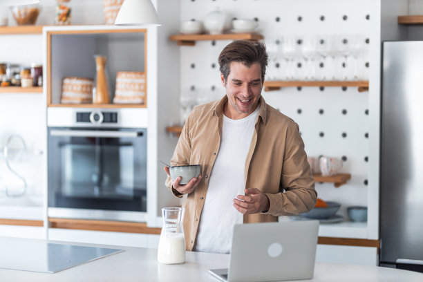 一个面带微笑的男性自由职业者在厨房里给自己做着牛奶早餐，用笔记本电脑工作。图片下载