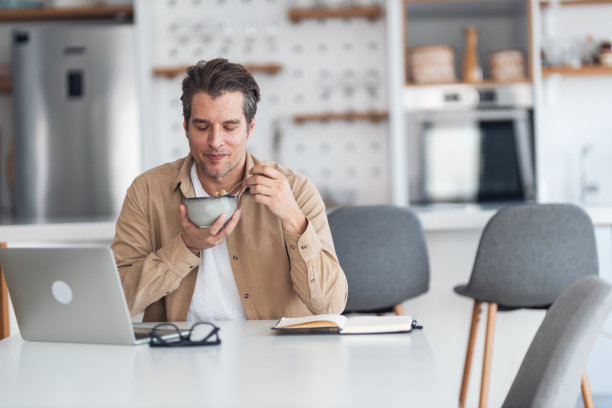 一名男性自由职业者一边吃早餐，一边在家里用笔记本电脑工作。图片下载