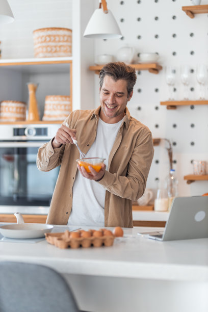 一位男性自由职业者一边用笔记本电脑工作，一边为早餐做鸡蛋。图片下载