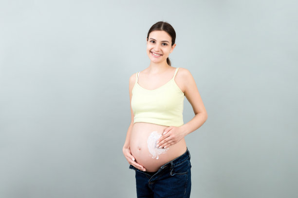 孕妇穿着敞开的牛仔裤，在腹部涂抹护肤霜，以防止妊娠纹，彩色背景与复制空间。健康肌肤理念图片下载