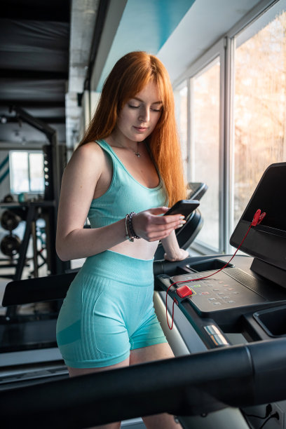 在健身房，年轻的红发女子站在跑步机教练旁边使用手机图片下载