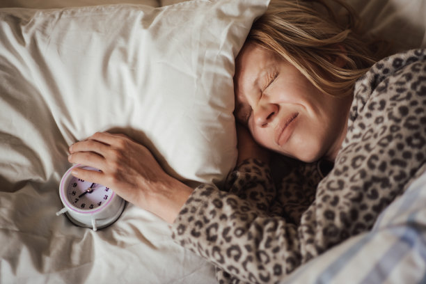 枕头上的闹钟和一个熟睡的女人，脸上带着不悦的表情。早上起床难，讨厌闹钟。睡眠问题的概念，失眠，昼夜节律。图片下载