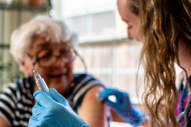 在前景中，一张特写照片显示，一位女护士正在给一位坐在餐桌旁的老妇人注射注射器。在后面，两人平静地坐着，护士安慰着他们。图片下载