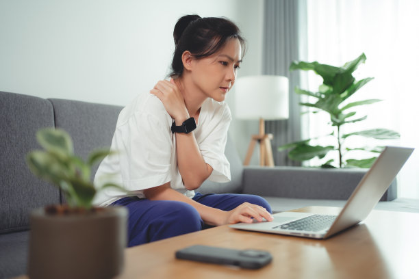 一名亚洲女性坐在家里的沙发上使用笔记本电脑，身体疼痛。图片下载
