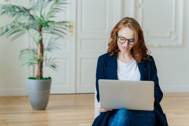 照片中，漂亮的姜黄色年轻女子戴着眼镜在网上观看教程视频，在现代公寓舒适的沙发上摆姿势，穿着优雅的衣服，享受免费的互联网连接图片下载