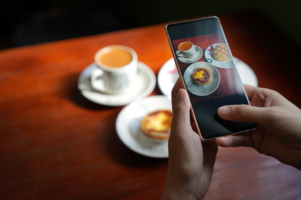从俯视的角度来看，一位年轻的女游客用她的智能手机拍下了美味的食物，港式马球包和蛋挞，还有一杯牛奶咖啡放在木桌上。图片下载