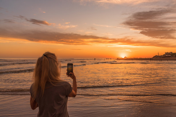 在海滩上用手机拍日落的女人。图片下载