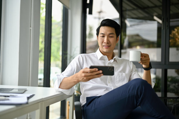 微笑的亚洲商人在手机上看视频，在办公桌前喝咖啡图片下载