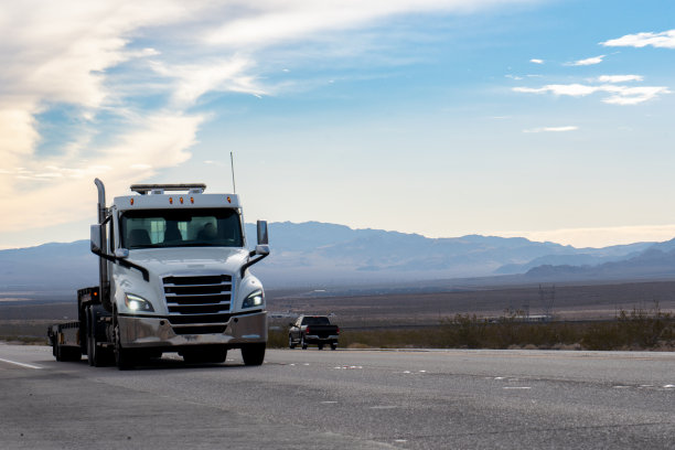 一辆白色的半挂车在广阔的内华达沙漠中行驶，夕阳西下，它的轮廓与雄伟的山脉并列，投射出温暖的金色光芒。图片下载