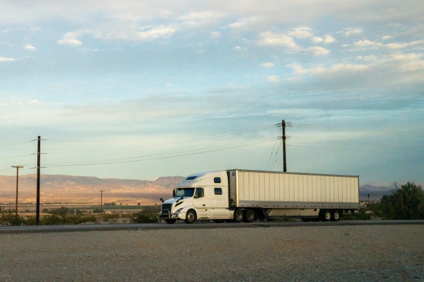 日落时分，一辆白色的半挂车在广阔的内华达沙漠中行驶，背后是雄伟的山脉。这景色是孤独与自然美景的和谐结合。图片下载