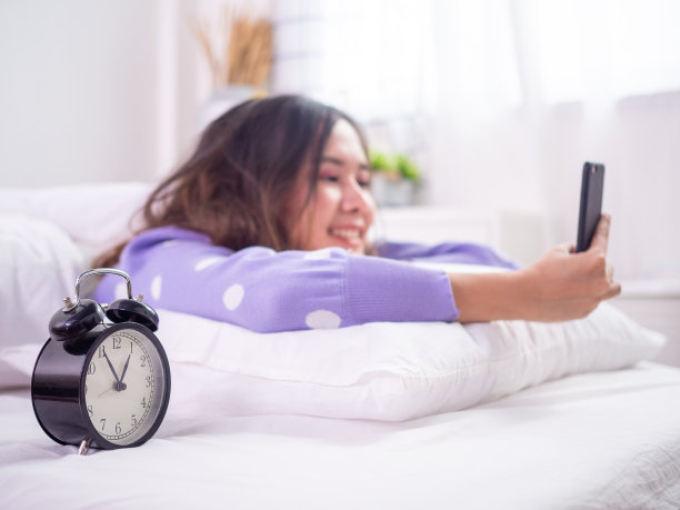 女人醒来后在床上使用电话。年轻的亚洲女性在床上玩游戏或愉快地聊天，闹钟显示时间。图片下载