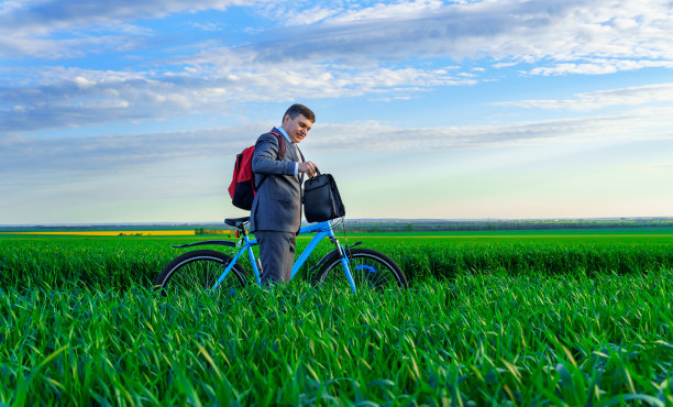 一位商人背着双肩包和公文包骑着自行车走在绿色的草地上，穿着西装，春天的美丽大自然，自由职业的商业理念图片下载