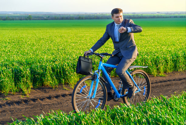 一个商人骑着自行车，拿着公文包，在绿色的草地上看手表，穿着西装，春天的美丽自然，自由职业的商业理念图片下载