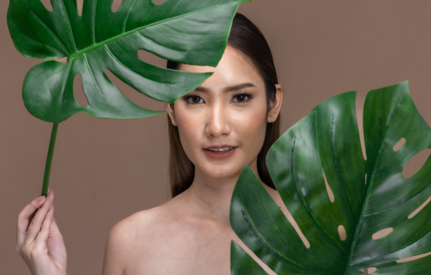 美丽的亚洲女人触摸眉毛微笑，干净和新鲜的皮肤幸福米色背景。美丽迷人的少女肖像与绿色植物的叶子隔绝。图片下载