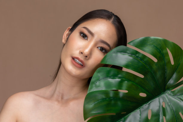 美丽的亚洲女人触摸眉毛微笑，干净和新鲜的皮肤幸福米色背景。美丽迷人的少女肖像与绿色植物的叶子隔绝。图片下载
