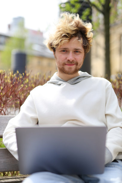 一个面带微笑的金发大胡子年轻人坐在长凳上，手里拿着一台笔记本电脑。图片下载