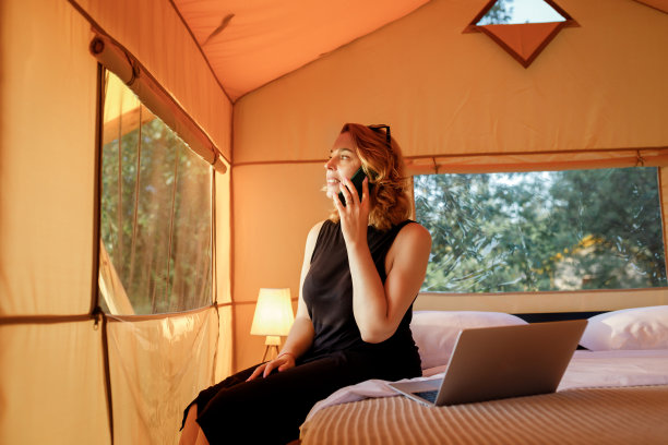 在一个阳光明媚的日子里，在舒适的豪华露营帐篷里工作时，微笑的自由职业者正在打电话。豪华户外露营帐篷，适合夏季度假度假。生活方式的概念图片下载