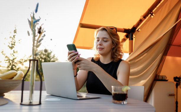 在一个阳光明媚的日子里，在舒适的豪华帐篷里用笔记本电脑工作时，微笑的自由职业者使用手机。豪华户外露营帐篷，适合夏季度假度假。生活方式的概念图片下载