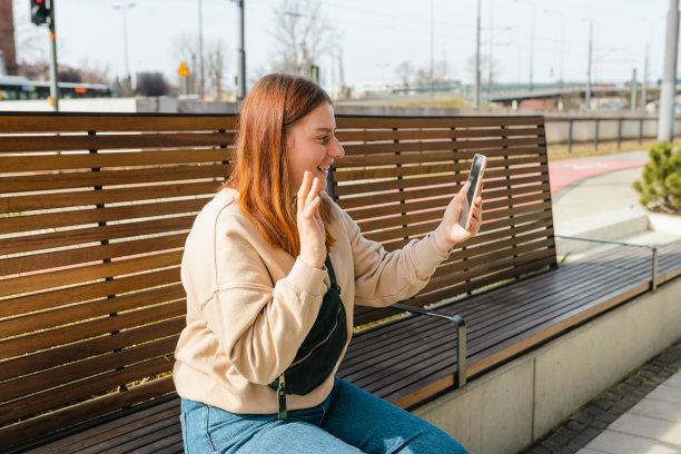 穿着便服的美女在城市街道上玩手机。年轻的潮人在城市背景下自拍。坐在长凳上在户外使用智能手机的人。图片下载