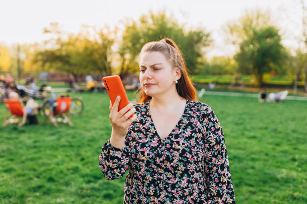 悲伤愤怒的女孩站在城市公园。消极的人的情绪，都市的生活方式观念。夏天，穿着裙子的年轻女子在公园户外使用智能手机。图片下载