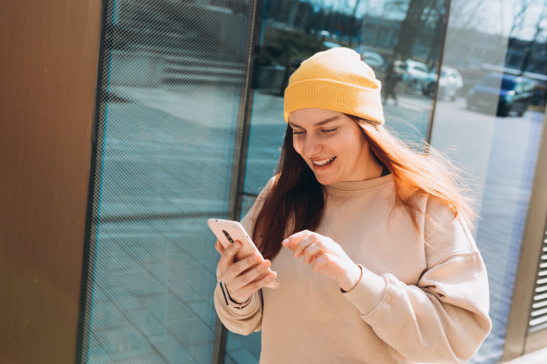 快乐开朗的年轻女子走在城市街道上查看她的智能手机。戴着黄色帽子的美女在户外使用智能手机的照片。城市生活方式概念。旅行者图片下载