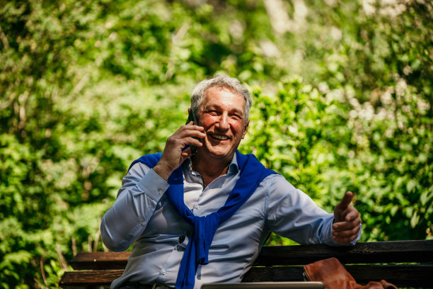 成熟的男人坐在长凳上，用电话在户外聊天。图片下载