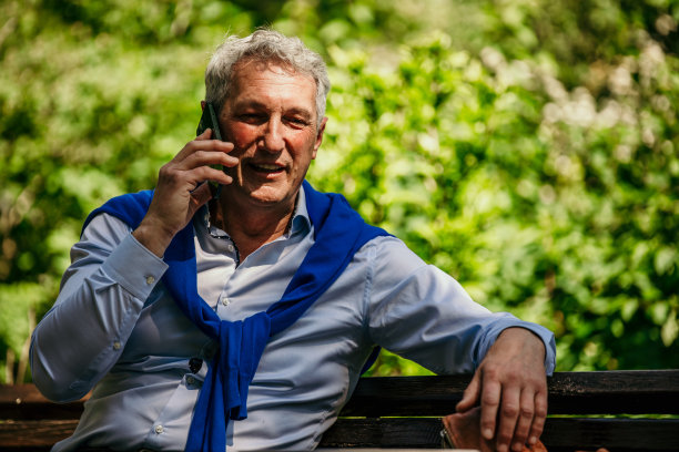 成熟的男人坐在长凳上，用电话在户外聊天。图片下载