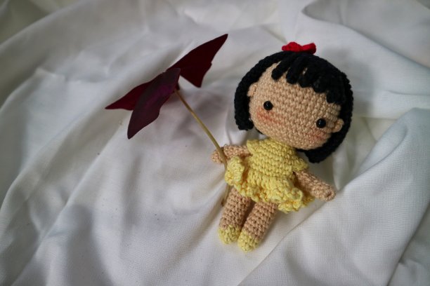 手工编织玩具，送给女婴的礼物，娃娃穿着黄裙子，头发上系着红蝴蝶结。留着黑色的短发，脸颊呈粉红色。钩针编织的漂亮女孩。图片下载