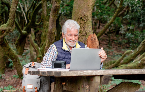 一位面带微笑的大胡子老人坐在公园户外的木桌前摆弄着笔记本电脑。智慧理念在大自然中工作，在新鲜空气中经商图片下载