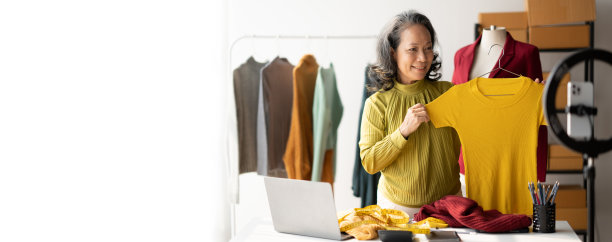 快乐的资深女性在网上做服装生意，测量，用智能手机在线为她的客户拿着漂亮的新衬衫。图片下载