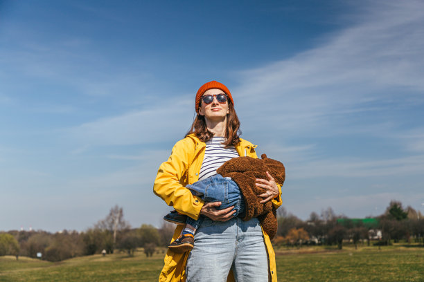 一位身穿黄色夹克、戴着红色帽子、戴着眼镜的母亲站在公园的草地上，以天空为背景，怀里抱着一个婴儿，正在哺乳，看起来像一个超级英雄图片下载