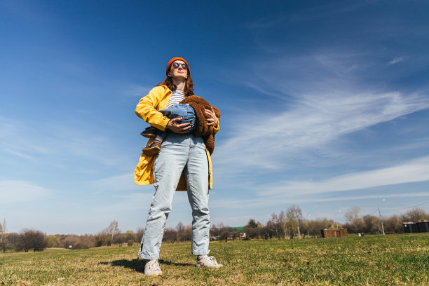 一位身穿黄色夹克、戴着红色帽子、戴着眼镜的母亲站在公园的草地上，以天空为背景，怀里抱着一个婴儿，正在哺乳，看起来像一个超级英雄图片下载