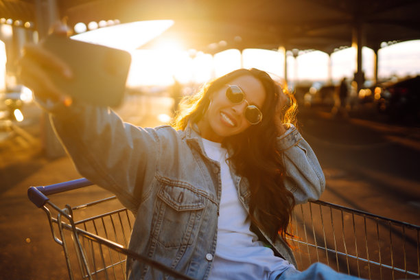 年轻女子拿着手机在购物车里开心地自拍。图片下载