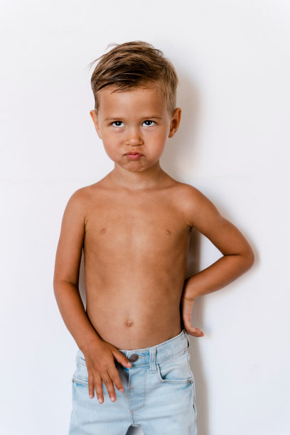 穿着蓝色牛仔裤的可爱小男孩在白色背景下摆姿势，脸上带着愚蠢的表情图片下载