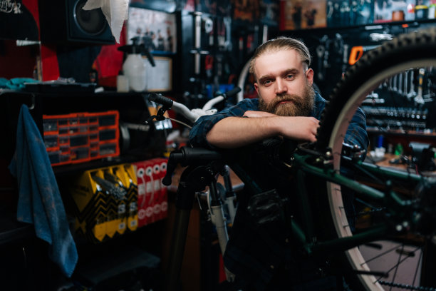 中镜头的肖像，英俊的自行车修理工有胡子站在自行车上靠在修理自行车车间黑暗的内部，看着相机。图片下载