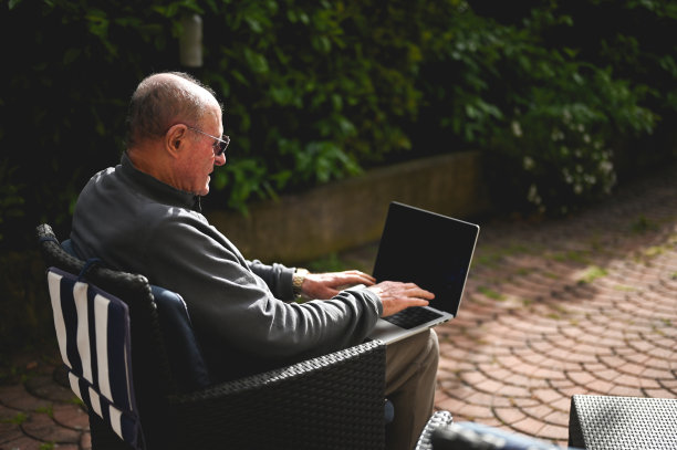 一位资深人士在正式的户外花园用笔记本电脑工作时的侧视图。图片下载