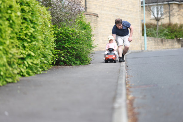 一个蹒跚学步的小女孩坐在爸爸推着的玩具车上，在一条住宅区的小路上疾驰而过。图片下载
