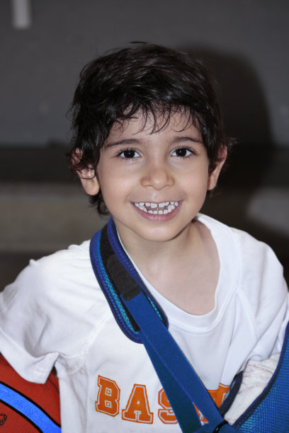 照片上，一个快乐而微笑的孩子，一只手打着石膏和吊带，另一只手拿着篮球。他正对着镜头。垂直人像摄影。有选择性的重点。本空间图片下载