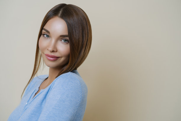 可爱的黑发女人的侧面照片与欧洲的外表，有健康的皮肤，精心护理，淡妆，穿着蓝色的套头衫，站在米色的背景，复制空间为您的推广图片下载