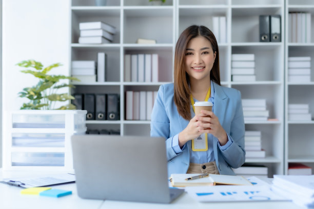 成功的亚洲女商人在办公室拿着咖啡杯，用笔记本电脑微笑。自信的亚洲女商人愉快地坐在办公室里。图片下载