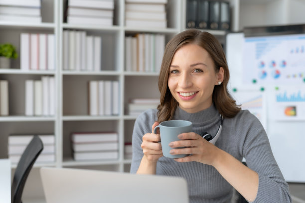 成功的女商人在办公室拿着咖啡杯，用笔记本电脑微笑。自信的女商人愉快地坐在办公室里。图片下载