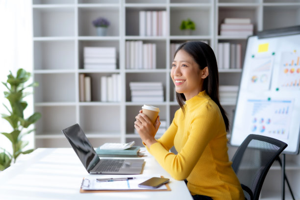 成功的亚洲女商人在办公室拿着咖啡杯，用笔记本电脑微笑。自信的亚洲女商人愉快地坐在办公室里。图片下载