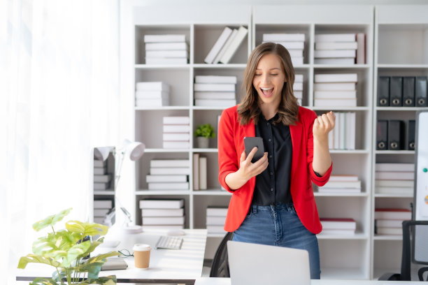快乐的女商人举起胜利的手，拿着智能手机开心地微笑。工作中成功的概念。图片下载