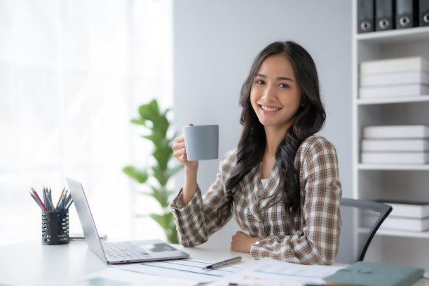 成功的亚洲女商人在办公室拿着咖啡杯，用笔记本电脑微笑。自信的亚洲女商人愉快地坐在办公室里。图片下载