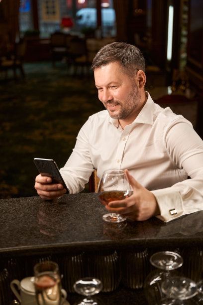 在酒吧里，一个微笑的男人拿着一杯白兰地在智能手机上浏览东西图片下载