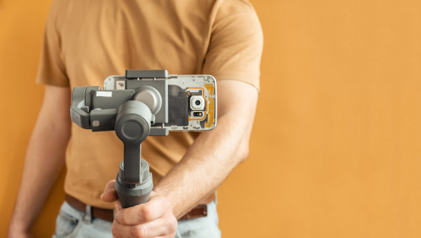 一名男子用智能手机在黄色背景上使用万向节稳定器进行拍摄。使用三轴电子稳定器进行vlog或视频拍摄。图片下载