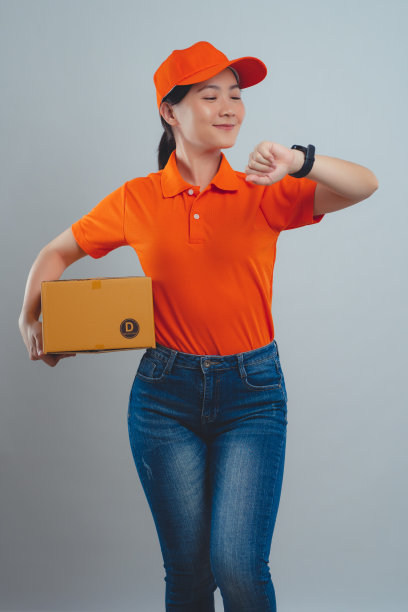 亚洲女人穿着统一的polo t恤，拿着箱子查看时间，很高兴能及时把包裹送到客户手中，孤立地站在白色的背景上。图片下载