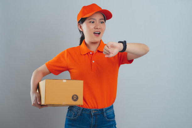 亚洲女人穿着统一的polo t恤，拿着盒子查看时间，因为给客户送包裹迟到而震惊，孤立地站在白色的背景上。图片下载