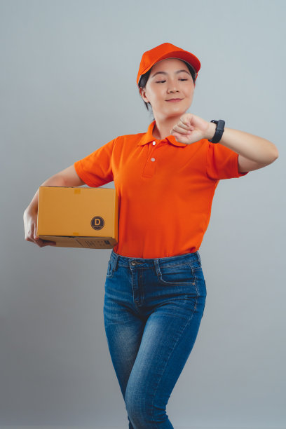 亚洲女人穿着统一的polo t恤，拿着箱子查看时间，很高兴能及时把包裹送到客户手中，孤立地站在白色的背景上。图片下载